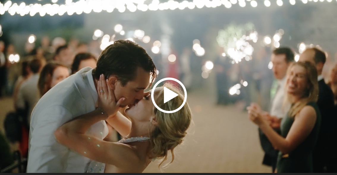 A couple shares a passionate kiss at their wedding, with the groom dipping the bride slightly. They are surrounded by guests holding sparklers, creating a magical and celebratory atmosphere with fairy lights visible in the background. Fearrington Village